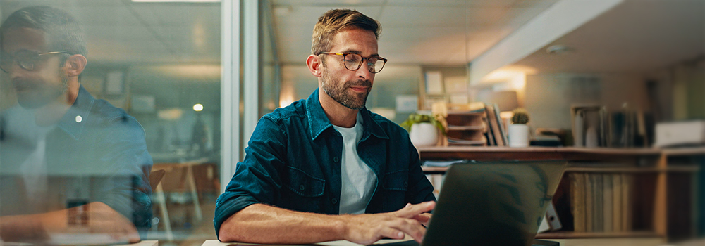 Person med briller og blå skjorte arbejder koncentreret på en bærbar computer i et moderne kontormiljø.