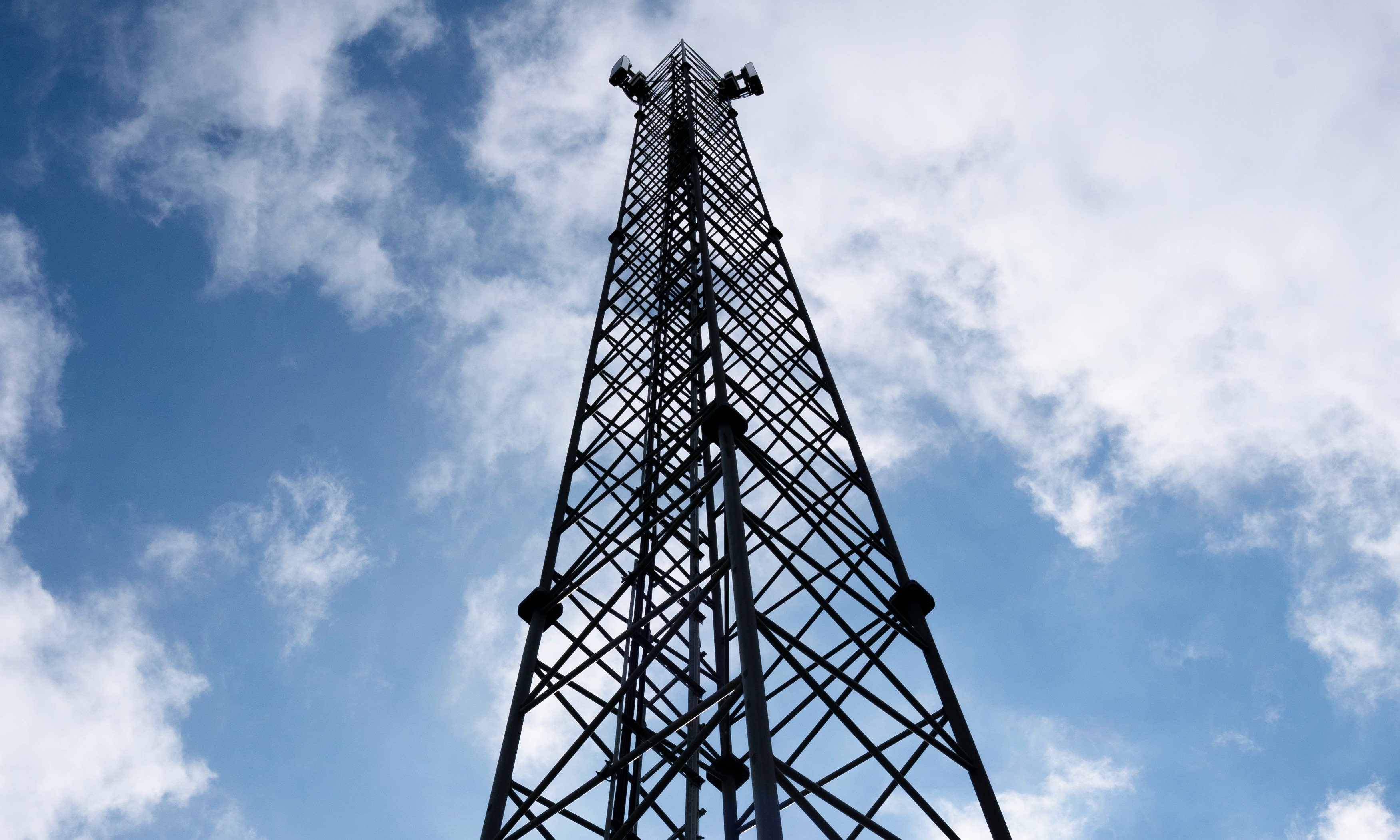 Høj telekommunikationsmast set nedefra mod en blå himmel med hvide skyer, silhuet af metalrammen rager op.