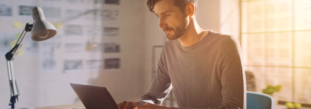 Person i grå sweater arbejder koncentreret på laptop ved skrivebord med lampe og notesbog i soloplyst hjemmekontor.