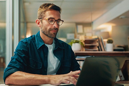 Person med briller og blå skjorte arbejder koncentreret på en bærbar computer i et lyst kontormiljø.