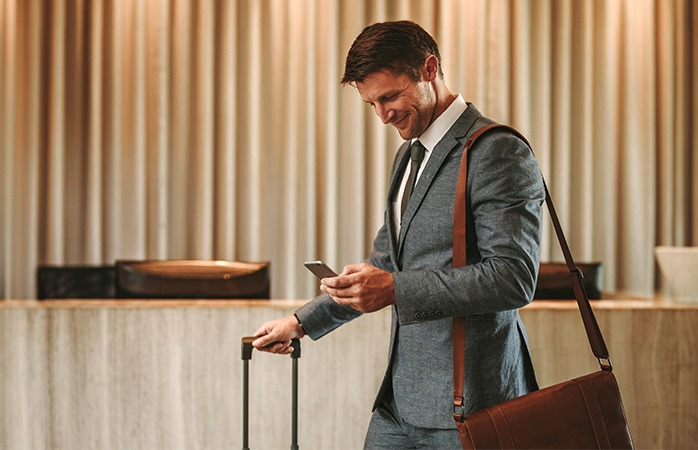 Forretningsmand i grå jakkesæt med brun taske tjekker sin telefon i en hotellobby, mens han holder en kuffert.