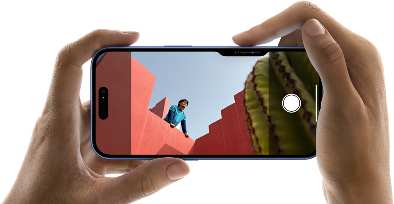 To hænder der holder en iPhone 16 vandret i hver deres ende, mobilens skærm er inde på kameraret, hvor man kan se en mand læne sig over solbrændt nuance murer, hvor manden kigger ud over murene foran ham, i højre hjørne kan man se en kaktus stikke frem 