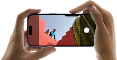 To hænder der holder en iPhone 16 vandret i hver deres ende, mobilens skærm er inde på kameraret, hvor man kan se en mand læne sig over solbrændt nuance murer, hvor manden kigger ud over murene foran ham, i højre hjørne kan man se en kaktus stikke frem