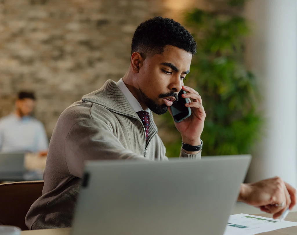 Person i beige sweater taler i telefon mens han arbejder på laptop i et kontor med murstensvæg og grønne planter.