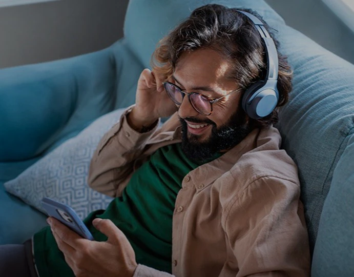 Person med skæg og briller sidder i blå sofa, lytter med blå hovedtelefoner og kigger på smartphone.