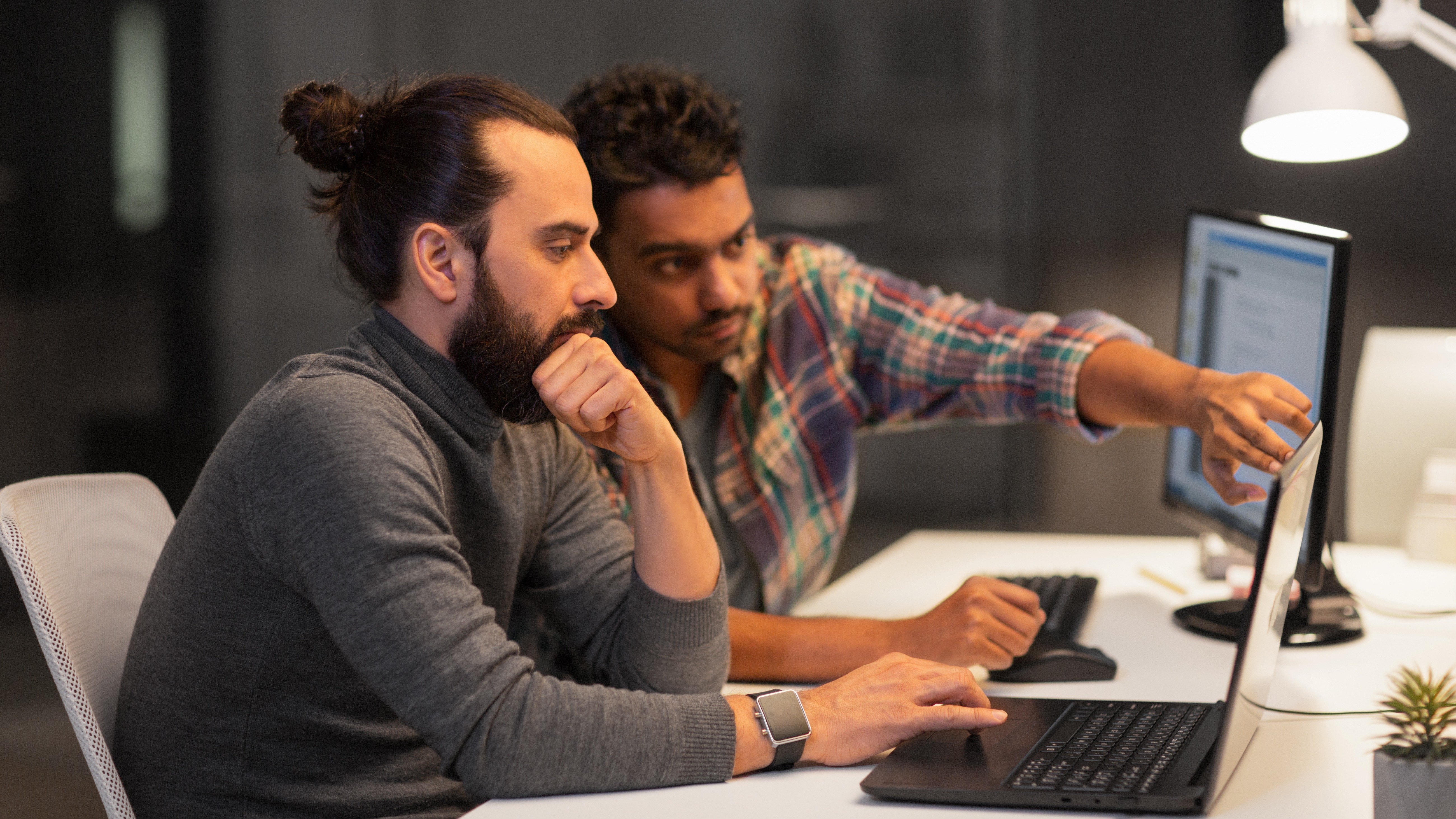 To personer arbejder koncentreret sammen ved computere i et mørkt kontor med en skrivebordslampe tændt.