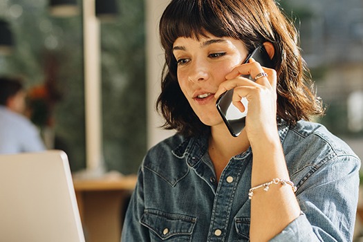 Kvinde i denimskjorte taler i telefon foran en bærbar computer i et lyst rum med naturligt lys.