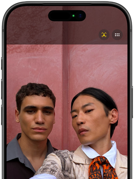 To personer tager en selfie foran en lyserød væg. Smartphone-skærm viser kameravisning med frontkamera aktiveret.