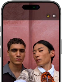 To personer tager en selfie foran en lyserød væg. Smartphone-skærm viser kameravisning med frontkamera aktiveret.