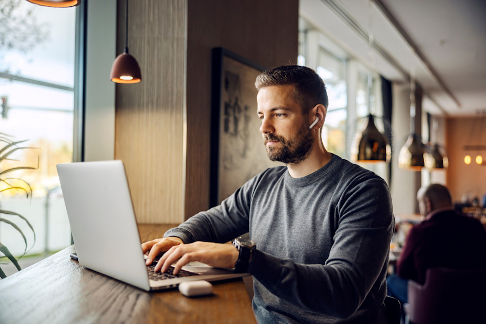 Mand i grå sweater arbejder på laptop ved vinduet i en café med trådløs høretelefon i øret og hængelamper over bordet.