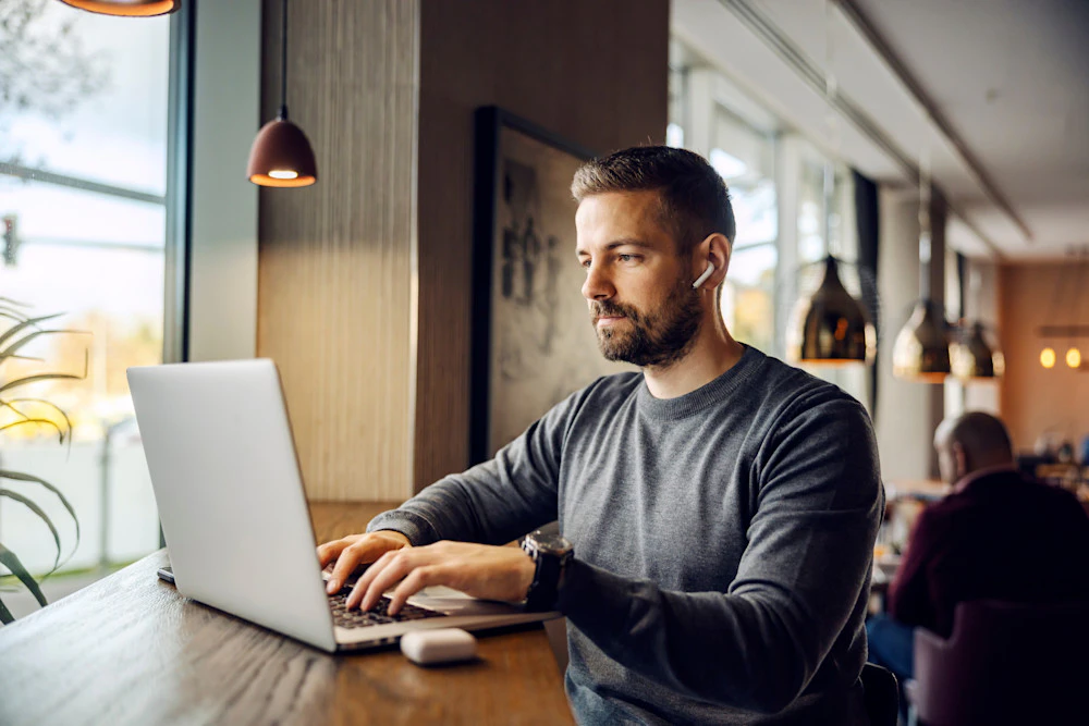 Mand i grå sweater arbejder på laptop ved vinduet i en café med trådløs høretelefon i øret og hængelamper over bordet.