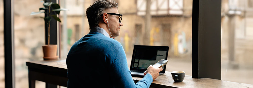 Mand sidder ved et cafébord. Han bruger en bærbar computer og holder en smartphone i hånden, mens han lytter til noget via høretelefoner.