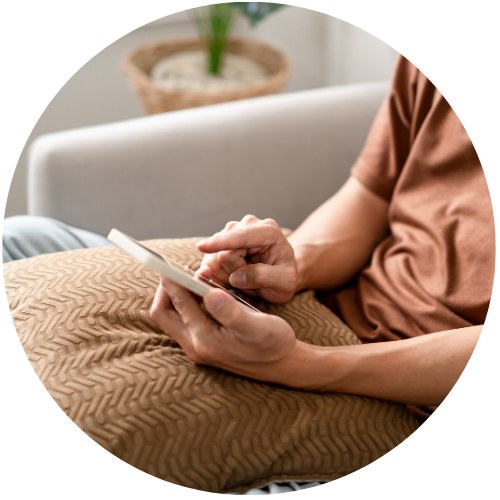 Person i brun trøje bruger smartphone på sofa med tekstureret beige pude i hyggelig stue med plante i baggrunden.