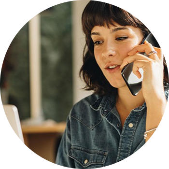 Kvinde i denimskjorte taler i telefon foran en bærbar computer i et lyst miljø med naturligt lys.