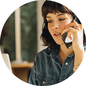 Kvinde i denimskjorte taler i telefon foran en bærbar computer i et lyst miljø med naturligt lys.