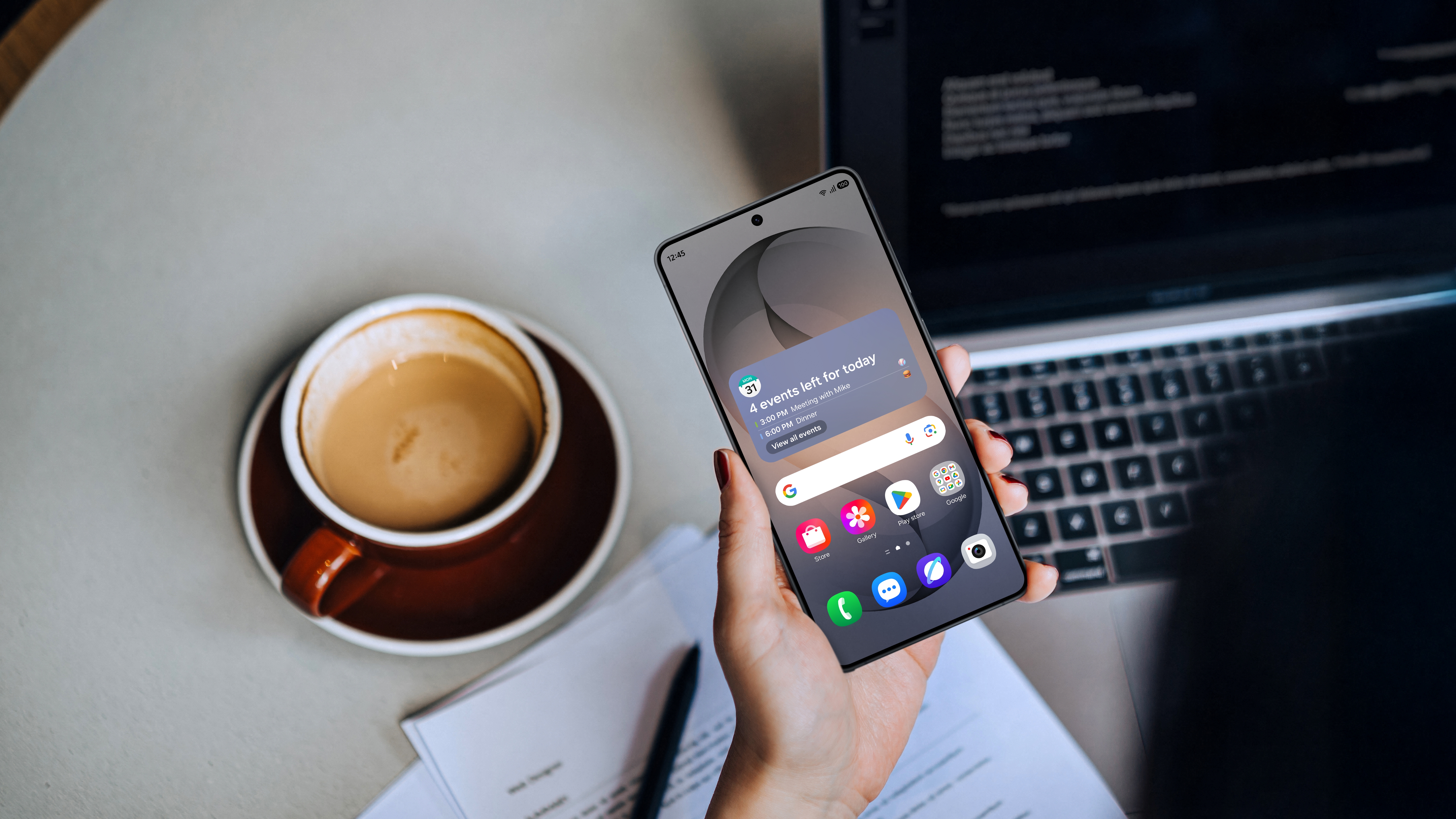 Hånd holder Samsung S26 smartphone med kalendernotifikationer ved siden af en kaffekop og laptop på et arbejdsbord.