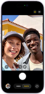 To smilende personer tager en selfie udendørs foran en gul bygning med rødt tag på en solrig dag.