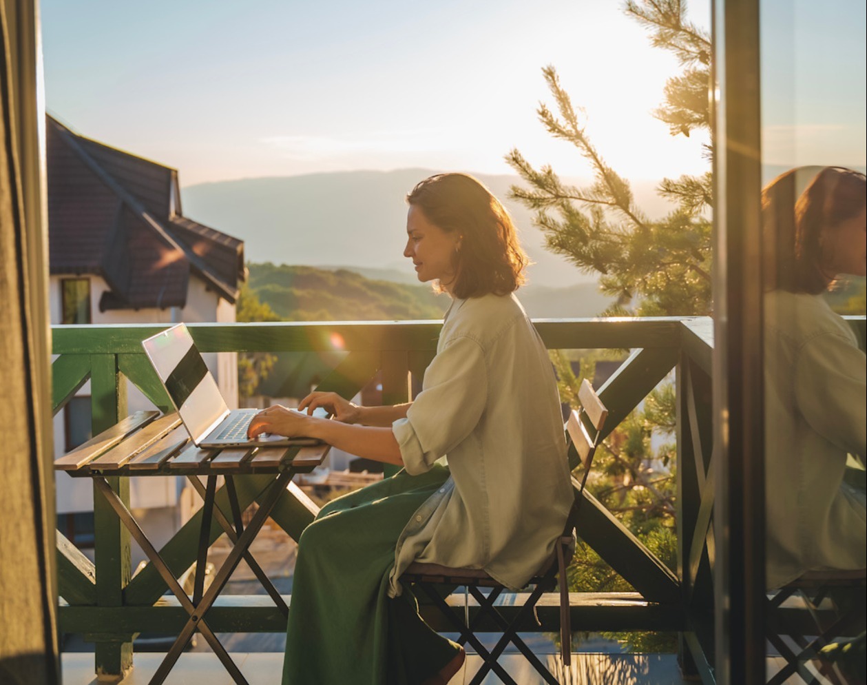 Kvinde arbejder på laptop på altan med bjerglandskab i baggrunden ved solnedgang, spejlet i vinduet.