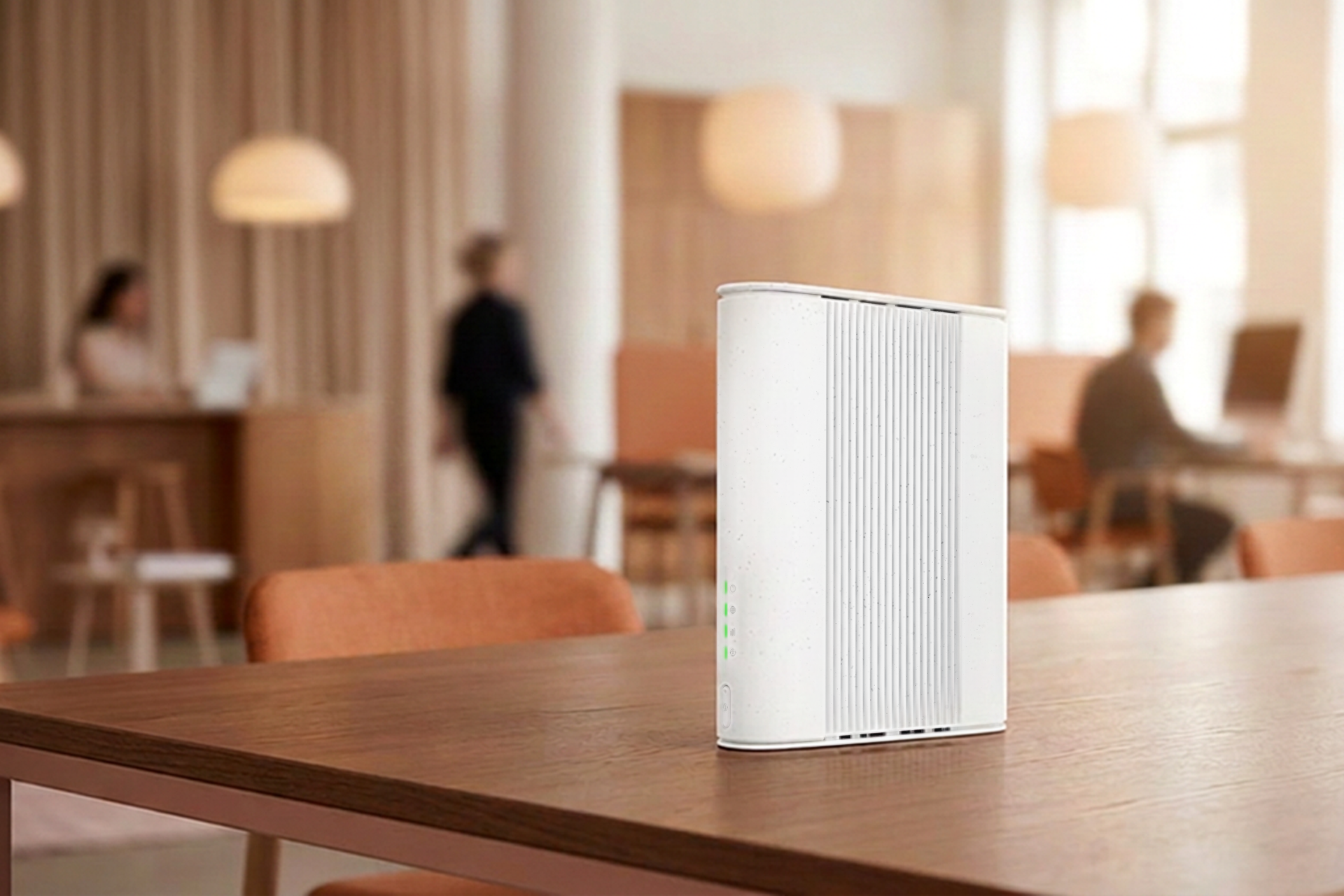 Hvid router med grønne LED-lys på et træbord i et lyst kontor med mennesker i baggrunden.