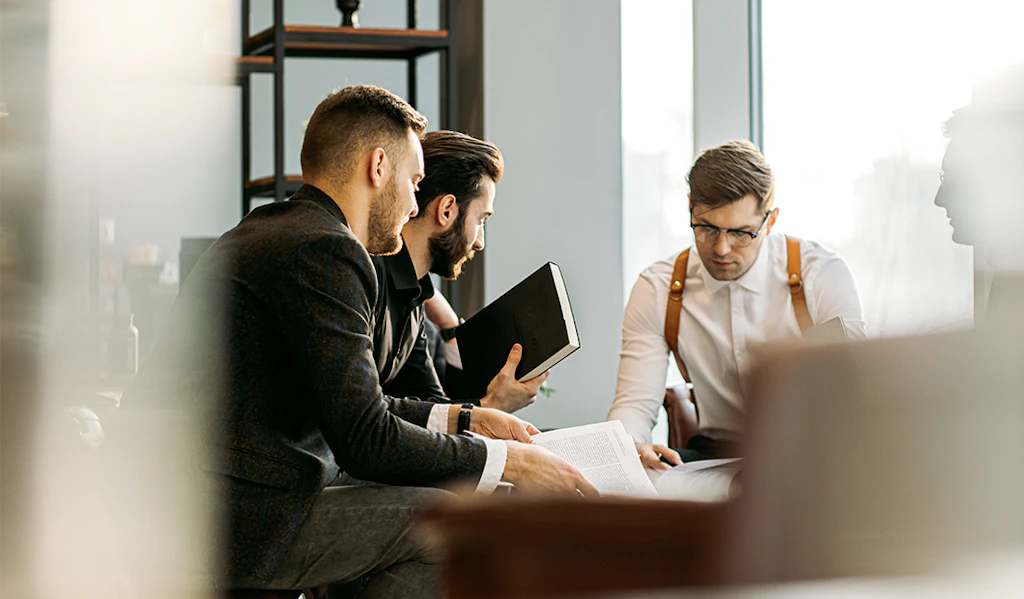Tre professionelle mænd i et lyst kontor samarbejder om dokumenter og en tablet ved et mødebord.