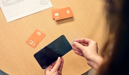 Person indsætter SIM-kort i smartphone. Orange SIM-kort ligger på træbord sammen med dokumenter.