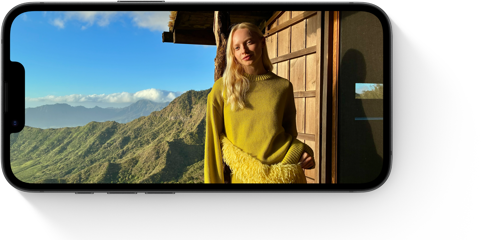 Person i gul sweater står ved træhytte med bjerglandskab i baggrunden, vist på en smartphone-skærm.