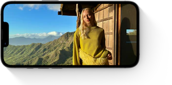 Person i gul sweater står ved træhytte med bjerglandskab i baggrunden, vist på en smartphone-skærm.