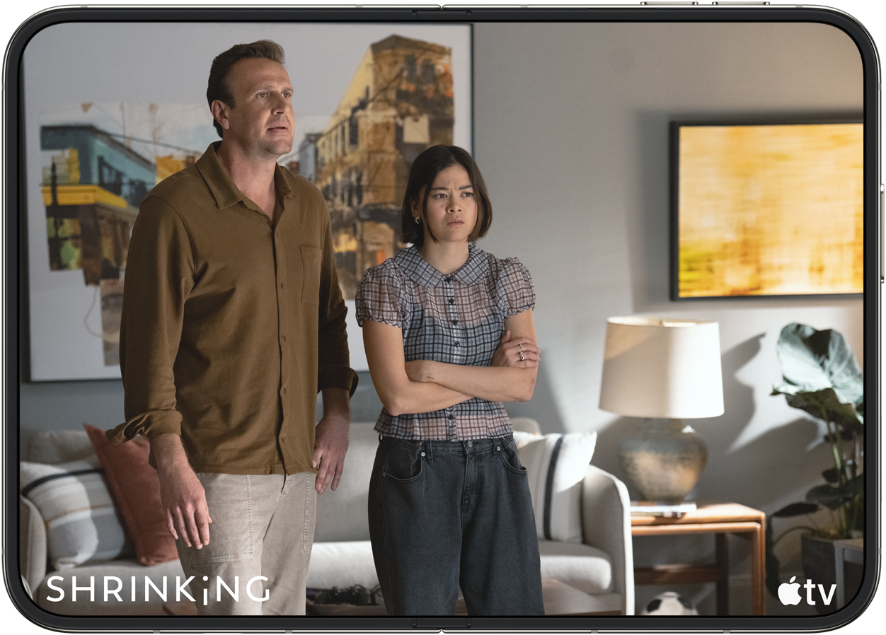 Scene fra Apple TV+ serien Shrinking med en mand i brun skjorte og en kvinde med korslagte arme i en stue.
