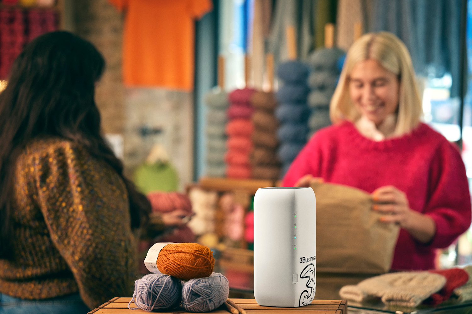 To kvinder i en garnbutik med en 3Business router i forgrunden og farverige garnnøgler på disken.