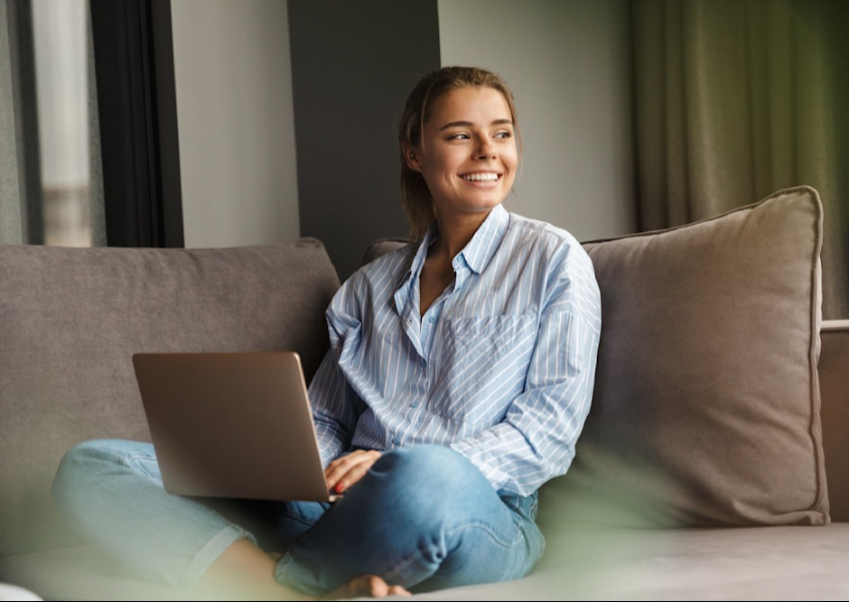 Smilende person i stribet skjorte sidder i sofa med bærbar computer, afslappet hjemmearbejde i lys stue.