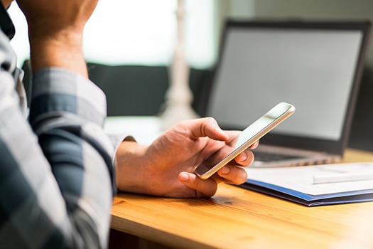 Person i ternet skjorte bruger smartphone ved skrivebord med laptop og kontorartikler i baggrunden.