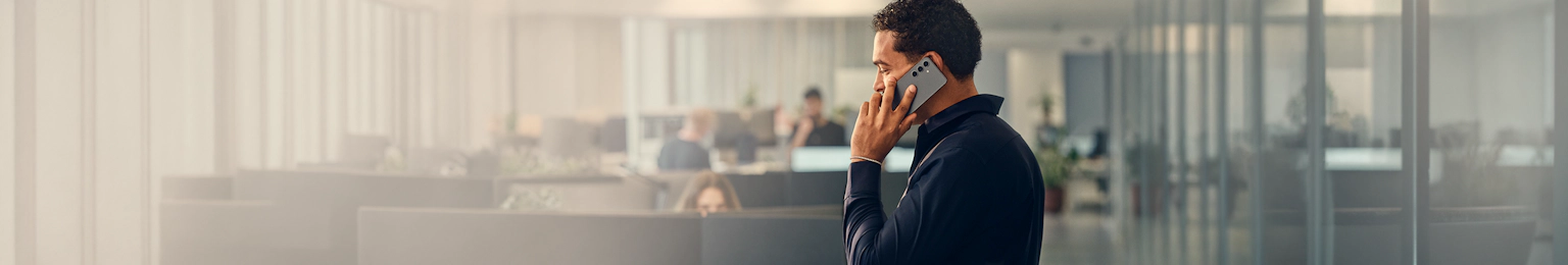 Person i mørkt tøj taler i mobiltelefon på et kontor med kolleger arbejdende i baggrunden.