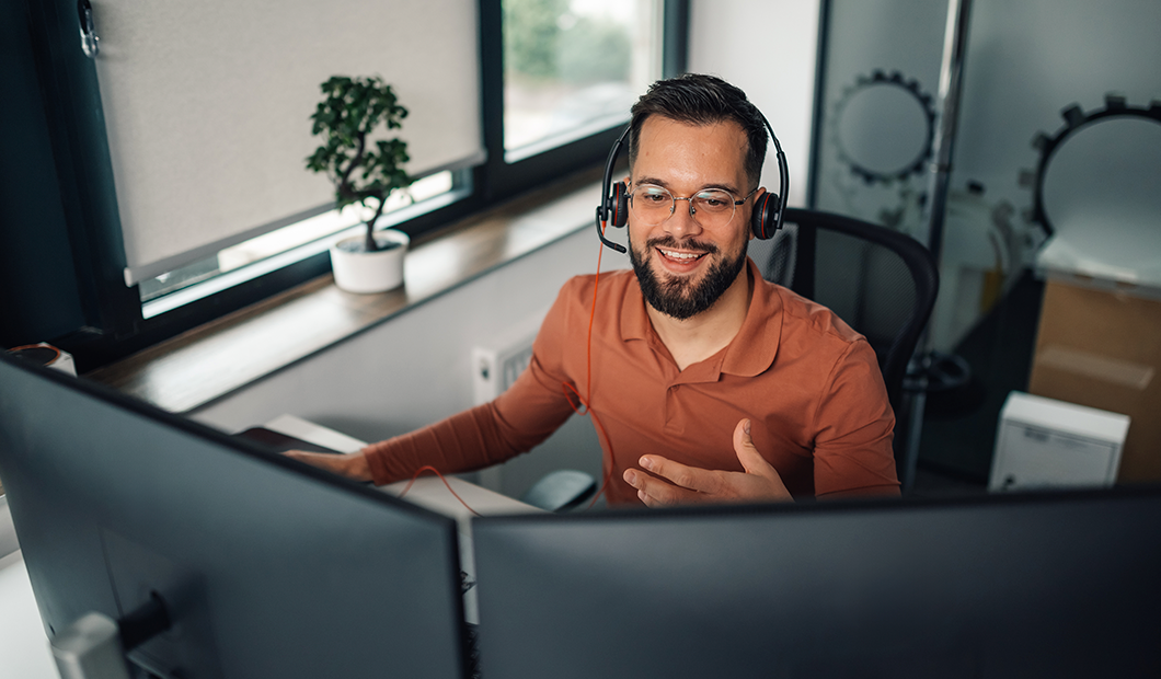 Person med headset og briller i orange skjorte smiler foran computerskærme i et kontor med vindue og lille plante.