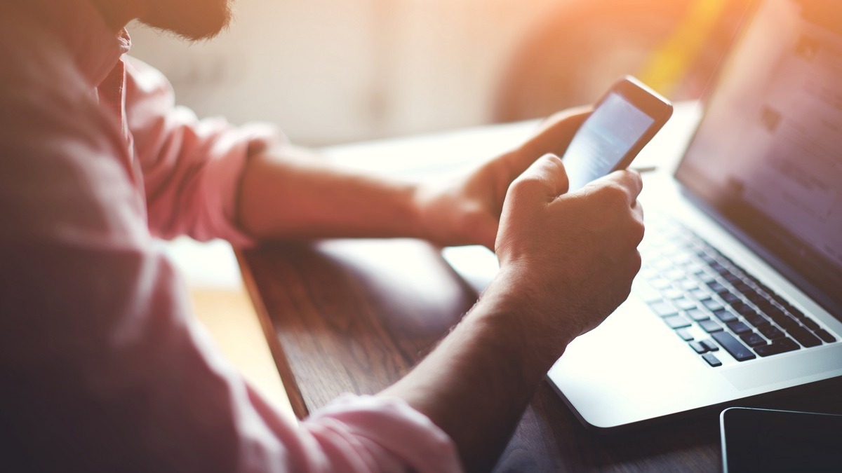 Person i lyserød skjorte tjekker smartphone ved siden af bærbar computer i varmt sollys ved træbord.