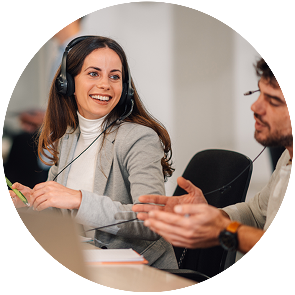 Smilende person med headset i grå blazer og hvid rullekrave sidder ved et møde med kolleger i et kontormiljø.