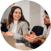 Smilende person med headset i grå blazer og hvid rullekrave sidder ved et møde med kolleger i et kontormiljø.