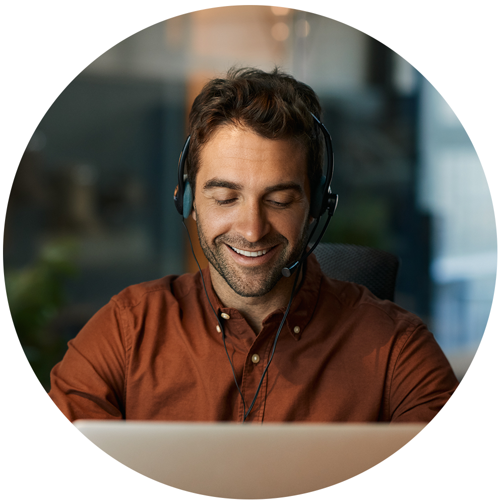 Person med headset smiler mens de arbejder på computer, iført brun skjorte i et kontor med sløret baggrund.