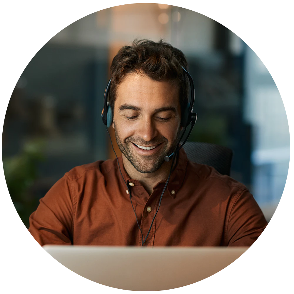 Person med headset smiler mens de arbejder på computer, iført brun skjorte i et kontor med sløret baggrund.