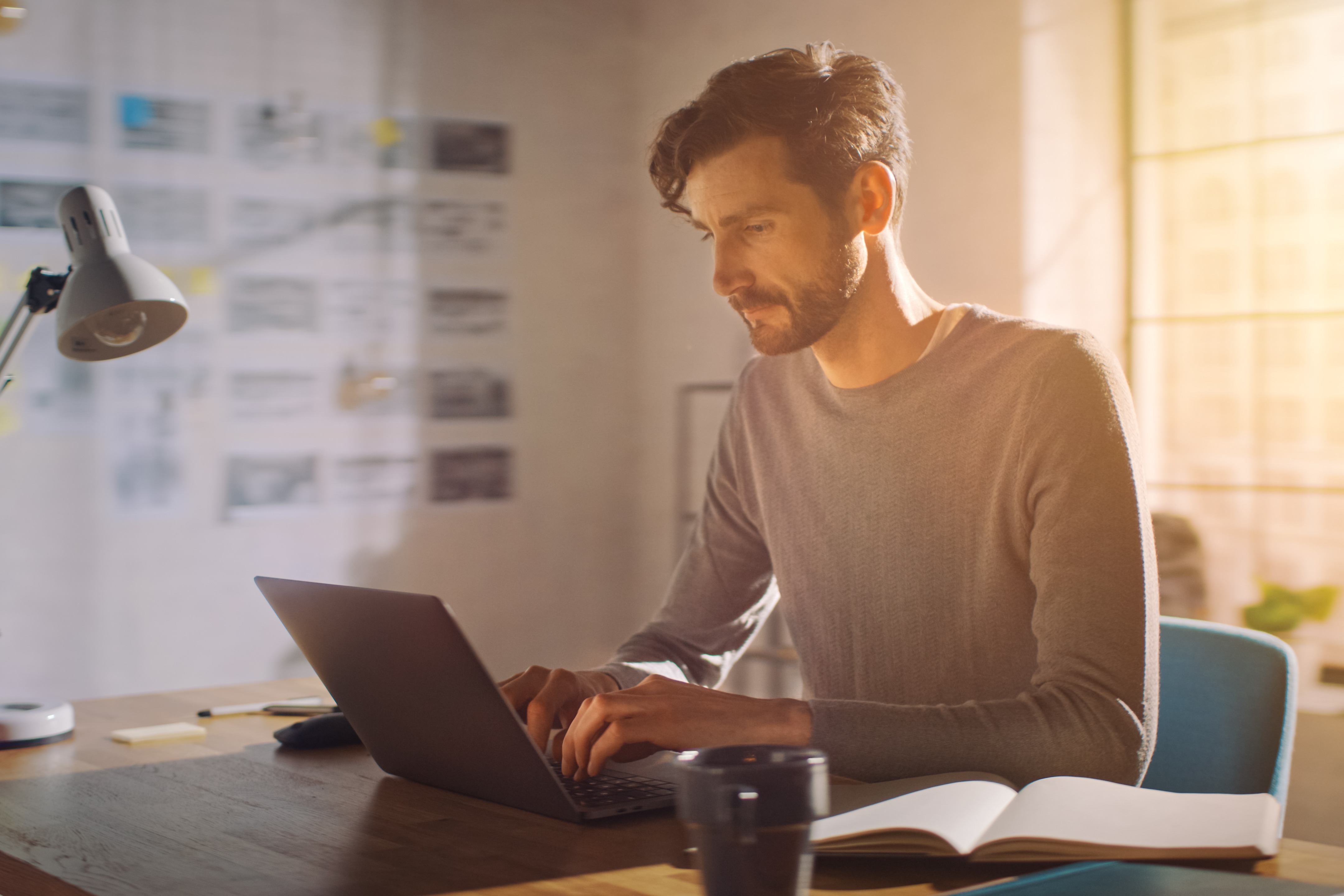 Person i grå sweater arbejder koncentreret på laptop ved skrivebord med notesbog og lampe i varmt sollys.