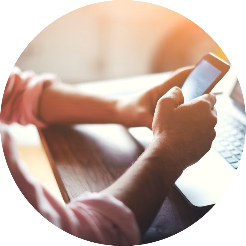 Person holder smartphone ved siden af en bærbar computer på et træbord i et lyst rum.