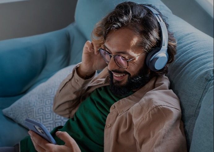 Person med skæg og briller sidder i blå sofa, iført høretelefoner og ser på smartphone med et smil.