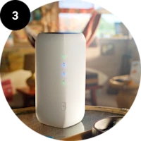 Hvid trådløs router med blå lysdioder står på et bord i et hjem. Nummer 3 vises i en sort cirkel.