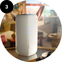 Hvid trådløs router med blå lysdioder står på et bord i et hjem. Nummer 3 vises i en sort cirkel.