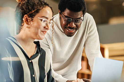 To personer med briller samarbejder ved en computer i et kontor med varm belysning.