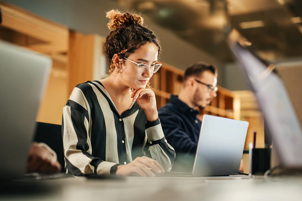 Person i blå blazer og briller bruger tablet i moderne café med træhylder og planter i baggrunden.