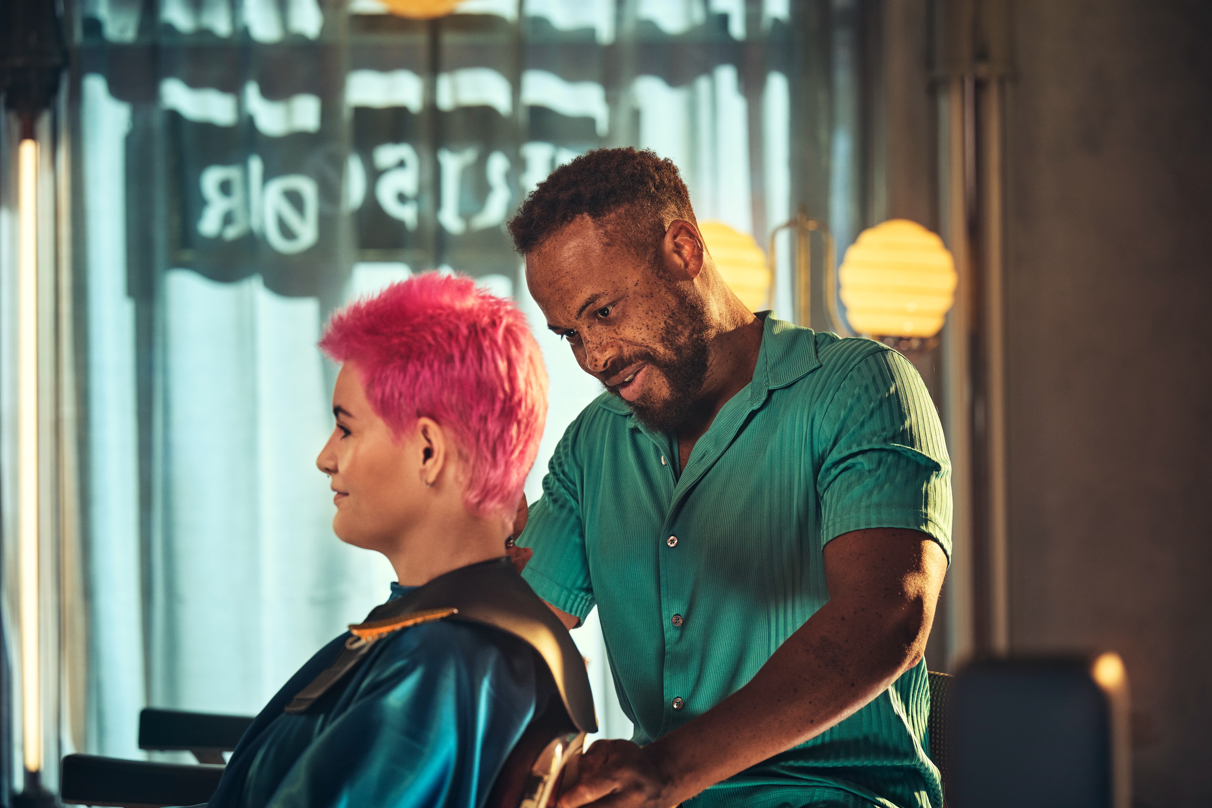 Person med lyserødt hår får frisørbehandling af en person i grøn skjorte i en salon med varmt lys.