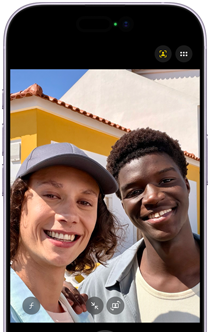 To personer smiler til kameraet foran en gul bygning med rødt tag under en lys himmel.