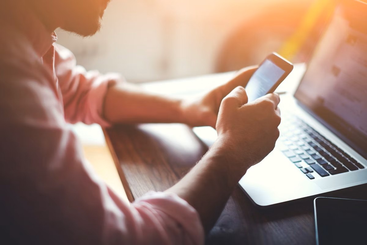 Person holder smartphone ved et træbord med en computer i nærheden, i et varmt, lyst rum.