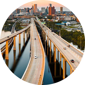 Luftfoto af motorvejsbroer over en flod med byens skyline i baggrunden ved solnedgang, badet i gyldent lys.