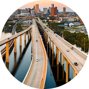 Luftfoto af motorvejsbroer over en flod med byens skyline i baggrunden ved solnedgang, badet i gyldent lys.